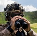 A military service member in desert MARPAT camouflage uniform is using binoculars to observe a distant area. The individual is wearing a tactical helmet with a camouflage cover, night vision device mount, and communications equipment. The setting appears to be a lush, green hilly or mountainous outdoor environment under partly cloudy skies.