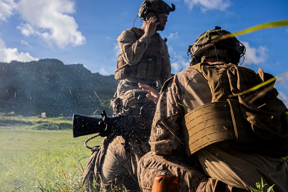 Two military personnel in full combat gear are positioned in a grassy field with mountainous terrain visible in the background. The foreground soldier appears to be firing a weapon, evidenced by visible debris/blast particles in the air, while the background soldier holds a communication device to his ear. Both wear camouflage uniforms, tactical vests, and helmets with mounted equipment.