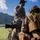 U.S. Marines with 3rd LCT participate in a live-fire basic skills package Two military personnel in full combat gear are positioned in a grassy field with mountainous terrain visible in the background. The foreground soldier appears to be firing a weapon, evidenced by visible debris/blast particles in the air, while the background soldier holds a communication device to his ear. Both wear camouflage uniforms, tactical vests, and helmets with mounted equipment.