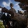 U.S. Marines with 3rd LCT participate in a live-fire basic skills package Military personnel in desert MARPAT camouflage uniforms are gathered around a crew-served weapon system, with one soldier pointing toward a target or direction of interest. The personnel are equipped with tactical gear including helmets with night-vision mounts, plate carriers, and sunglasses. The setting appears to be an outdoor training or operational environment with vegetation and a partly cloudy sky visible.