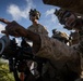 Military personnel in desert MARPAT camouflage uniforms are gathered around a crew-served weapon system, with one soldier pointing toward a target or direction of interest. The personnel are equipped with tactical gear including helmets with night-vision mounts, plate carriers, and sunglasses. The setting appears to be an outdoor training or operational environment with vegetation and a partly cloudy sky visible.