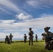 A group of military personnel in full combat gear are positioned across an open grassy field overlooking a coastal body of water. The foreground soldier is kneeling with a rifle at the ready, while others stand in dispersed formation behind. The setting appears to be a tropical or subtropical coastal location based on the lush vegetation and ocean backdrop.