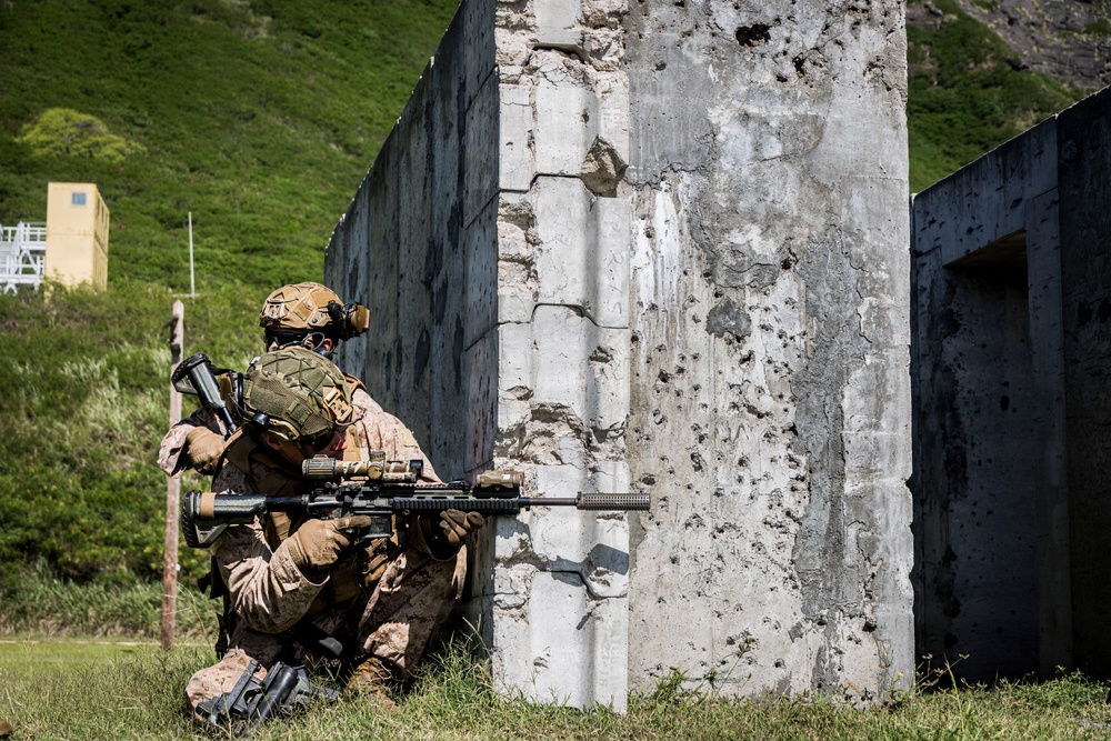 Two military personnel in desert camouflage uniforms are positioned at the corner of a concrete wall, with one aiming a rifle in a tactical shooting stance. The setting appears to be a military training range, evidenced by the bullet-pocked concrete structures and controlled environment. The soldiers are equipped with full combat gear including tactical helmets, body armor, and optical accessories.