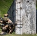 Two military personnel in desert camouflage uniforms are positioned at the corner of a concrete wall, with one aiming a rifle in a tactical shooting stance. The setting appears to be a military training range, evidenced by the bullet-pocked concrete structures and controlled environment. The soldiers are equipped with full combat gear including tactical helmets, body armor, and optical accessories.