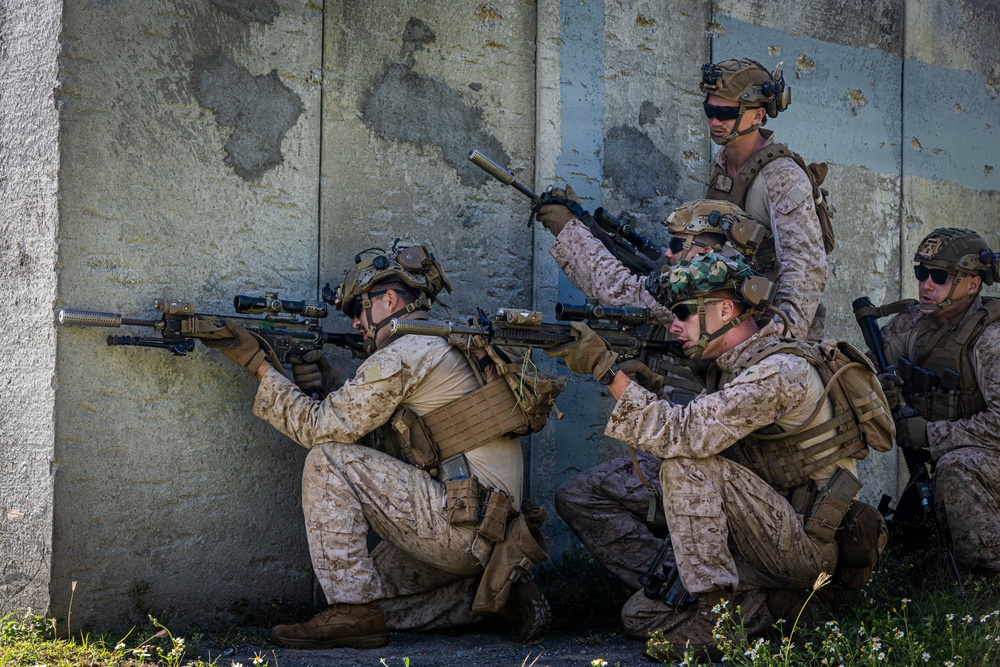 Four military personnel in desert camouflage uniforms are positioned against a concrete wall in a tactical formation. Two soldiers in the foreground are aiming rifles around a corner while others provide overwatch and security. The scene appears to be a military training exercise or tactical operation.