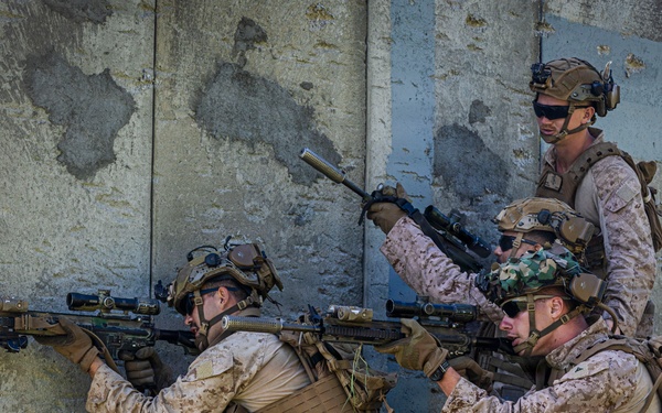 Four military personnel in desert camouflage uniforms are positioned against a concrete wall in a tactical formation. Two soldiers in the foreground are aiming rifles around a corner while others provide overwatch and security. The scene appears to be a military training exercise or tactical operation.