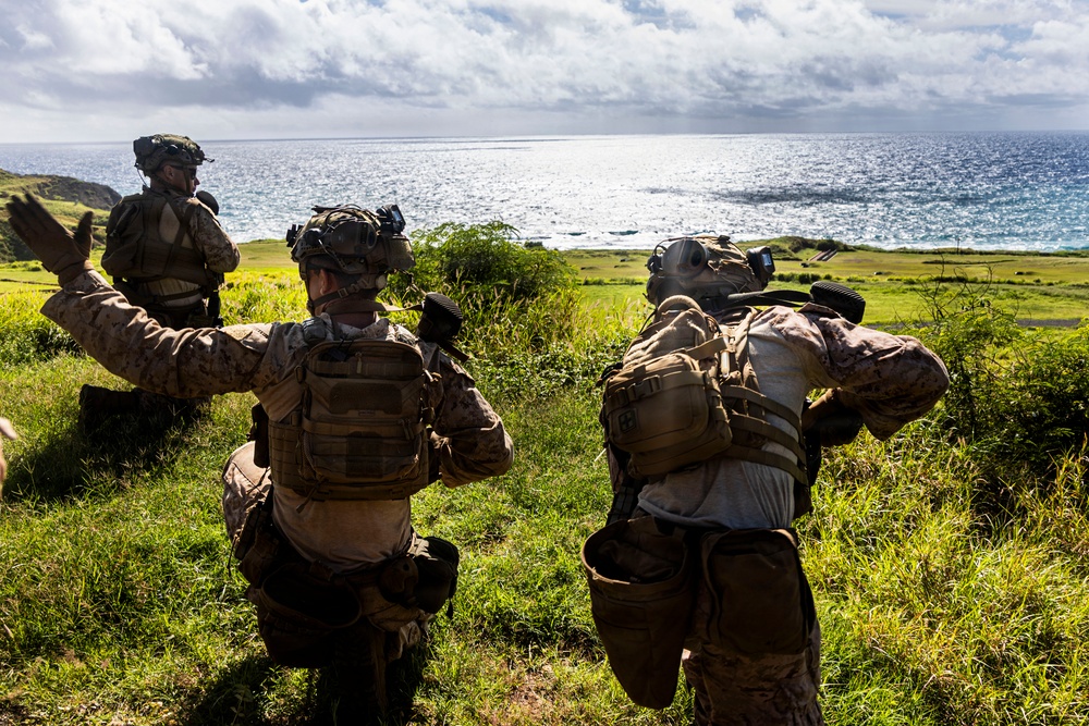Three military personnel in full tactical gear are positioned on a grassy coastal hillside, appearing to conduct a field exercise or reconnaissance operation. One soldier is giving a hand signal with arm raised, suggesting active communication or coordination. The scene overlooks a coastal area with green terrain and ocean visible in the background.