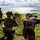 U.S. Marines with 3rd LCT participate in a live-fire basic skills package Three military personnel in full tactical gear are positioned on a grassy coastal hillside, appearing to conduct a field exercise or reconnaissance operation. One soldier is giving a hand signal with arm raised, suggesting active communication or coordination. The scene overlooks a coastal area with green terrain and ocean visible in the background.