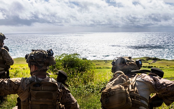 Three military personnel in full tactical gear are positioned on a grassy coastal hillside, appearing to conduct a field exercise or reconnaissance operation. One soldier is giving a hand signal with arm raised, suggesting active communication or coordination. The scene overlooks a coastal area with green terrain and ocean visible in the background.