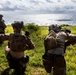 Three military personnel in full tactical gear are positioned on a grassy coastal hillside, appearing to conduct a field exercise or reconnaissance operation. One soldier is giving a hand signal with arm raised, suggesting active communication or coordination. The scene overlooks a coastal area with green terrain and ocean visible in the background.