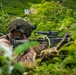 A soldier in full tactical gear is positioned prone in dense vegetation, aiming a rifle equipped with an optical scope. The individual is wearing desert camouflage uniform, a tactical helmet with accessories, and protective gloves. The setting appears to be a lush, green outdoor environment, consistent with a training exercise or field operation.