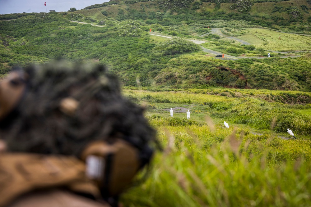 A military or law enforcement training exercise is taking place at an outdoor shooting range set in a lush, hilly green landscape. A shooter in the foreground, wearing a camouflage helmet and tactical gear, is aiming downrange at two white humanoid silhouette targets positioned in the field. The range appears to be a live-fire facility with safety markers, roads, and a red flag visible in the background.