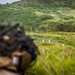 A military or law enforcement training exercise is taking place at an outdoor shooting range set in a lush, hilly green landscape. A shooter in the foreground, wearing a camouflage helmet and tactical gear, is aiming downrange at two white humanoid silhouette targets positioned in the field. The range appears to be a live-fire facility with safety markers, roads, and a red flag visible in the background.