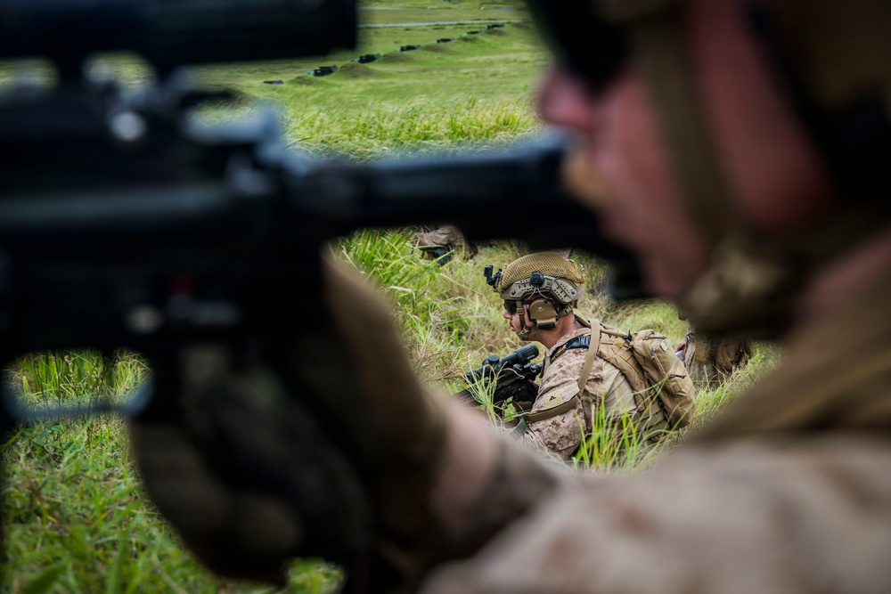 The image shows military personnel in tactical field positions on a grassy terrain, likely during a training exercise. Soldiers wearing desert camouflage uniforms and tactical helmets with mounted equipment are positioned low in the grass. The foreground features a soldier aiming a rifle, with additional personnel visible in the middle ground.