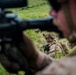 The image shows military personnel in tactical field positions on a grassy terrain, likely during a training exercise. Soldiers wearing desert camouflage uniforms and tactical helmets with mounted equipment are positioned low in the grass. The foreground features a soldier aiming a rifle, with additional personnel visible in the middle ground.