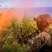 A soldier in full combat gear is positioned in dense vegetation near a coastline, appearing to be in a surveillance or overwatch posture. The individual is equipped with a tactical helmet with mounted accessories, camouflage uniform, and is holding a firearm. The image is captured during golden hour with a warm lens flare effect, with the ocean visible in the background.