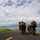 U.S. Marines with 3rd LCT participate in a live-fire basic skills package A column of military personnel in full combat gear marches along a paved road toward a coastal landscape with mountains and turquoise water in the background. The soldiers are carrying rifles and large backpacks, with at least one individual shouldering what appears to be a rocket launcher or similar crew-served weapon. The setting suggests a tropical or Pacific island environment, consistent with a training exercise or patrol.