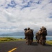A column of military personnel in full combat gear marches along a paved road toward a coastal landscape with mountains and turquoise water in the background. The soldiers are carrying rifles and large backpacks, with at least one individual shouldering what appears to be a rocket launcher or similar crew-served weapon. The setting suggests a tropical or Pacific island environment, consistent with a training exercise or patrol.