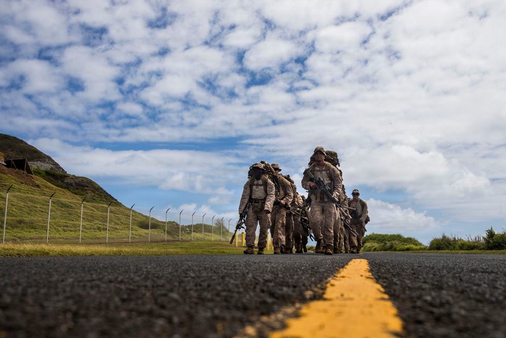 A low-angle ground-level photograph captures a group of military personnel in camouflage uniforms marching along a paved road with double yellow center lines. The soldiers are carrying full combat gear including rifles and large rucksacks, suggesting a tactical road march or training exercise. The setting appears to be a military installation with green hills, a perimeter fence, and a partly cloudy sky in the background.