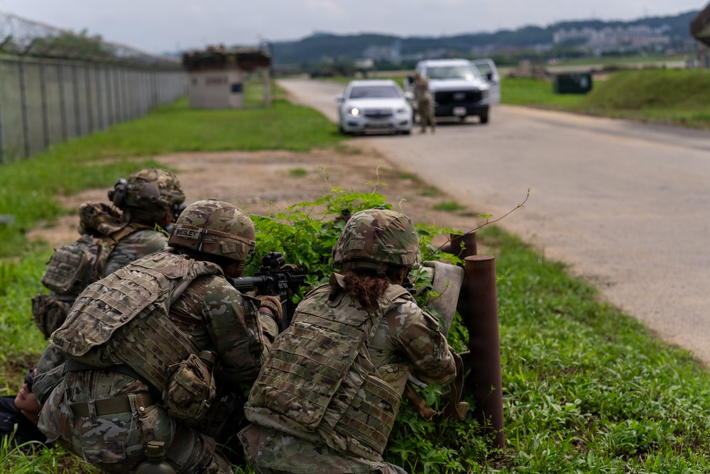 Military personnel in full combat gear and camouflage uniforms are taking covered positions behind vegetation along a road, with weapons aimed toward vehicles stopped ahead. A separate individual in uniform is visible near the vehicles in the background, suggesting a checkpoint or vehicle control point scenario. The scene appears to be a military training exercise based on the controlled environment and base infrastructure visible.