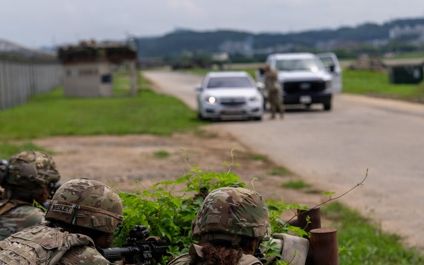 Military personnel in full combat gear and camouflage uniforms are taking covered positions behind vegetation along a road, with weapons aimed toward vehicles stopped ahead. A separate individual in uniform is visible near the vehicles in the background, suggesting a checkpoint or vehicle control point scenario. The scene appears to be a military training exercise based on the controlled environment and base infrastructure visible.