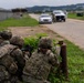 Military personnel in full combat gear and camouflage uniforms are taking covered positions behind vegetation along a road, with weapons aimed toward vehicles stopped ahead. A separate individual in uniform is visible near the vehicles in the background, suggesting a checkpoint or vehicle control point scenario. The scene appears to be a military training exercise based on the controlled environment and base infrastructure visible.
