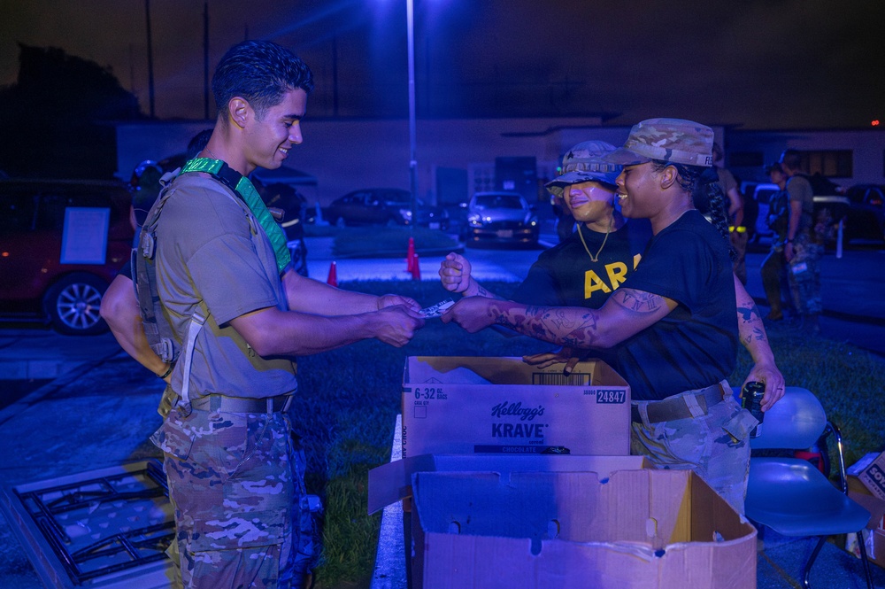 A nighttime military scene showing personnel in Army uniforms and tactical gear exchanging items near cardboard boxes. The setting appears to be an outdoor distribution or checkpoint station, illuminated by artificial blue-tinted lighting. Three primary individuals are visible, with one wearing a green sash/identifier and tactical vest interacting with two others in Army PT-style black shirts and camouflage caps.
