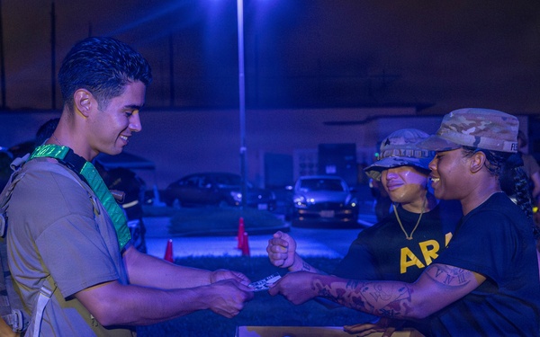 A nighttime military scene showing personnel in Army uniforms and tactical gear exchanging items near cardboard boxes. The setting appears to be an outdoor distribution or checkpoint station, illuminated by artificial blue-tinted lighting. Three primary individuals are visible, with one wearing a green sash/identifier and tactical vest interacting with two others in Army PT-style black shirts and camouflage caps.