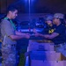 A nighttime military scene showing personnel in Army uniforms and tactical gear exchanging items near cardboard boxes. The setting appears to be an outdoor distribution or checkpoint station, illuminated by artificial blue-tinted lighting. Three primary individuals are visible, with one wearing a green sash/identifier and tactical vest interacting with two others in Army PT-style black shirts and camouflage caps.