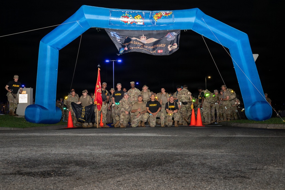 A group of military personnel, primarily U.S. Army soldiers in camouflage uniforms, pose for a group photo under a blue inflatable arch at night. A banner hanging from the arch reads 'Royal Danish Air Force March' and 'Veterans March,' indicating a multinational military marching event. Several soldiers are wearing black 'ARMY' t-shirts, and a red unit guidon flag is visible among the group.