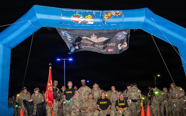 A group of military personnel, primarily U.S. Army soldiers in camouflage uniforms, pose for a group photo under a blue inflatable arch at night. A banner hanging from the arch reads 'Royal Danish Air Force March' and 'Veterans March,' indicating a multinational military marching event. Several soldiers are wearing black 'ARMY' t-shirts, and a red unit guidon flag is visible among the group.