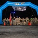 A group of military personnel, primarily U.S. Army soldiers in camouflage uniforms, pose for a group photo under a blue inflatable arch at night. A banner hanging from the arch reads 'Royal Danish Air Force March' and 'Veterans March,' indicating a multinational military marching event. Several soldiers are wearing black 'ARMY' t-shirts, and a red unit guidon flag is visible among the group.