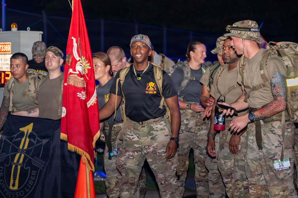 A group of military personnel in camouflage uniforms are gathered at night, likely at the start or finish of a ruck march or endurance event. A prominent red unit guidon flag is visible, along with a black Special Forces-style flag. The central figure wears a black shirt with '1-4 ADA BN' (1st Battalion, 4th Air Defense Artillery) insignia.