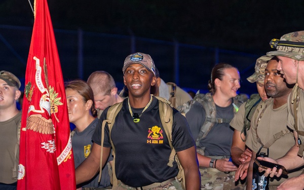 A group of military personnel in camouflage uniforms are gathered at night, likely at the start or finish of a ruck march or endurance event. A prominent red unit guidon flag is visible, along with a black Special Forces-style flag. The central figure wears a black shirt with '1-4 ADA BN' (1st Battalion, 4th Air Defense Artillery) insignia.