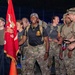 A group of military personnel in camouflage uniforms are gathered at night, likely at the start or finish of a ruck march or endurance event. A prominent red unit guidon flag is visible, along with a black Special Forces-style flag. The central figure wears a black shirt with '1-4 ADA BN' (1st Battalion, 4th Air Defense Artillery) insignia.