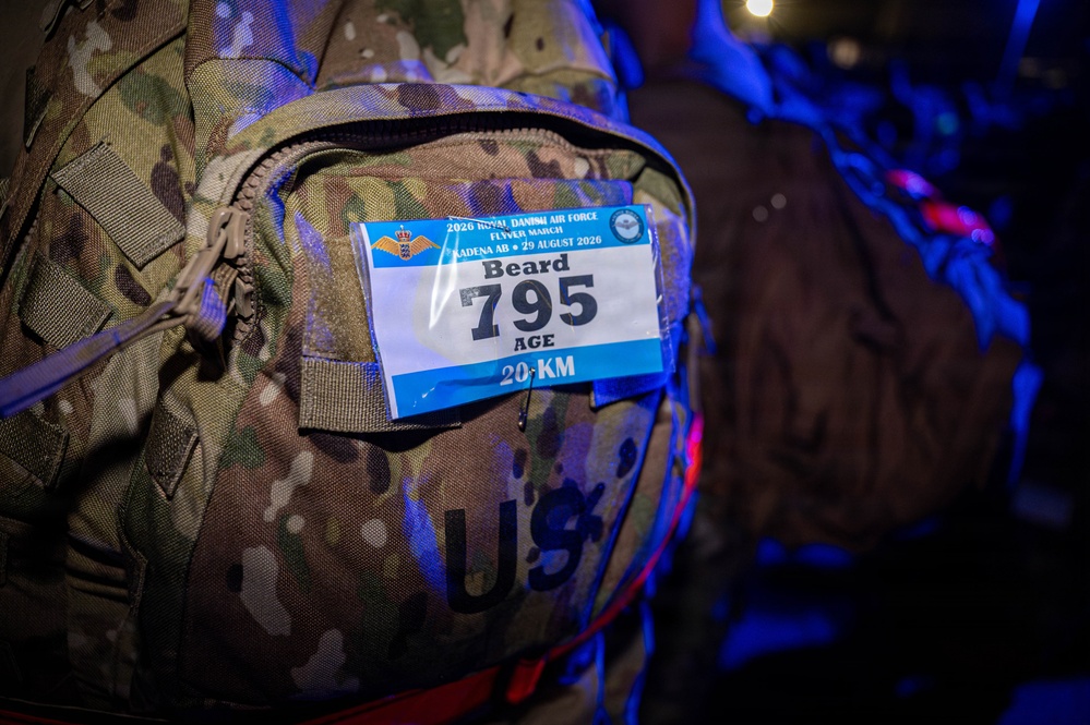 A close-up photograph of a military camouflage backpack bearing a race bib attached to its exterior. The bib is from the 2026 Royal Danish Air Force Flyer March event held at Kadena AB on 29 August 2026, assigned to participant 'Beard' with bib number 795 for the 20 KM distance. The backpack is marked with 'U.S.' indicating it belongs to a U.S. military service member.