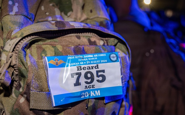 A close-up photograph of a military camouflage backpack bearing a race bib attached to its exterior. The bib is from the 2026 Royal Danish Air Force Flyer March event held at Kadena AB on 29 August 2026, assigned to participant 'Beard' with bib number 795 for the 20 KM distance. The backpack is marked with 'U.S.' indicating it belongs to a U.S. military service member.