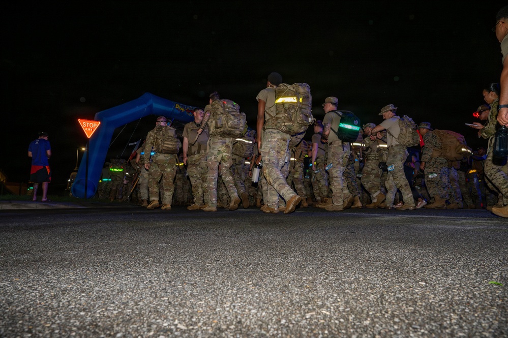 A group of military personnel in camouflage uniforms and full rucksacks are marching at night through what appears to be a ruck march or endurance event. They are passing under or near a blue inflatable arch, suggesting an organized start/finish line for a military fitness or memorial event. Reflective gear and safety lighting are visible on participants.