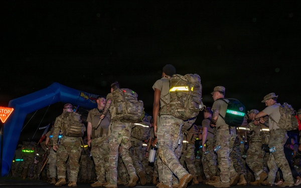 A group of military personnel in camouflage uniforms and full rucksacks are marching at night through what appears to be a ruck march or endurance event. They are passing under or near a blue inflatable arch, suggesting an organized start/finish line for a military fitness or memorial event. Reflective gear and safety lighting are visible on participants.