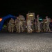 A group of military personnel in camouflage uniforms and full rucksacks are marching at night through what appears to be a ruck march or endurance event. They are passing under or near a blue inflatable arch, suggesting an organized start/finish line for a military fitness or memorial event. Reflective gear and safety lighting are visible on participants.
