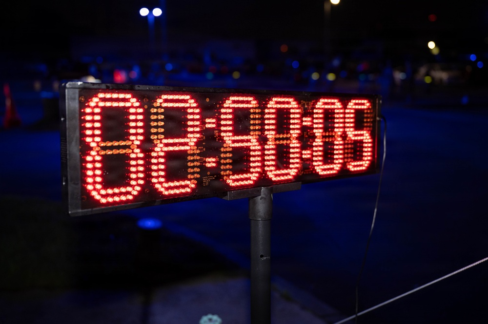 A large LED digital timer/clock display is mounted on a pole stand, photographed at night during what appears to be an outdoor event. The display shows red LED digits reading approximately '88:60:08' or a similar time format, though some digits may be mid-transition. The background shows a dark outdoor environment with scattered colored lights in the bokeh.
