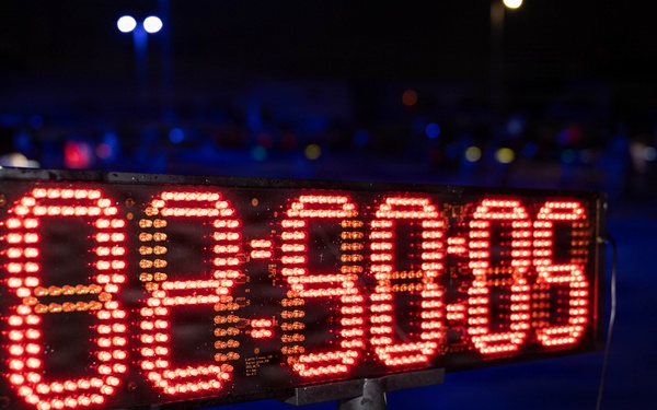 A large LED digital timer/clock display is mounted on a pole stand, photographed at night during what appears to be an outdoor event. The display shows red LED digits reading approximately '88:60:08' or a similar time format, though some digits may be mid-transition. The background shows a dark outdoor environment with scattered colored lights in the bokeh.