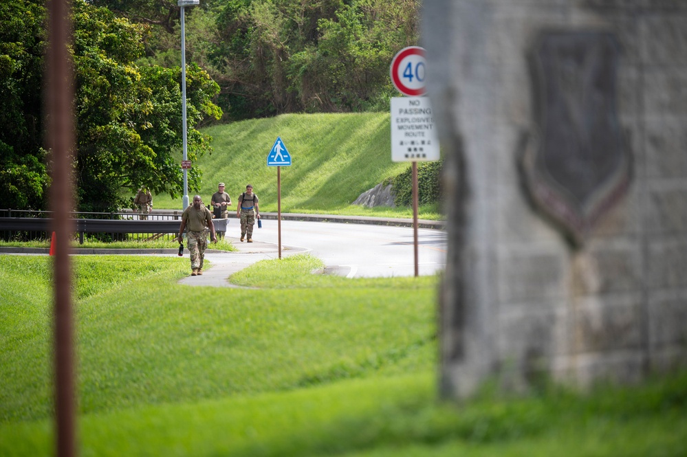 The image shows military personnel in camouflage uniforms walking along a sidewalk path on what appears to be a military installation. Road signs are visible including a 40 km/h speed limit sign and a 'No Passing - Explosives Movement Route' sign with bilingual text (English and Japanese/Okinawan characters). A blurred military unit insignia or emblem is visible on a stone structure in the foreground right.