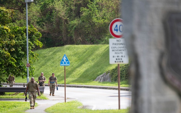 The image shows military personnel in camouflage uniforms walking along a sidewalk path on what appears to be a military installation. Road signs are visible including a 40 km/h speed limit sign and a 'No Passing - Explosives Movement Route' sign with bilingual text (English and Japanese/Okinawan characters). A blurred military unit insignia or emblem is visible on a stone structure in the foreground right.