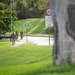 The image shows military personnel in camouflage uniforms walking along a sidewalk path on what appears to be a military installation. Road signs are visible including a 40 km/h speed limit sign and a 'No Passing - Explosives Movement Route' sign with bilingual text (English and Japanese/Okinawan characters). A blurred military unit insignia or emblem is visible on a stone structure in the foreground right.