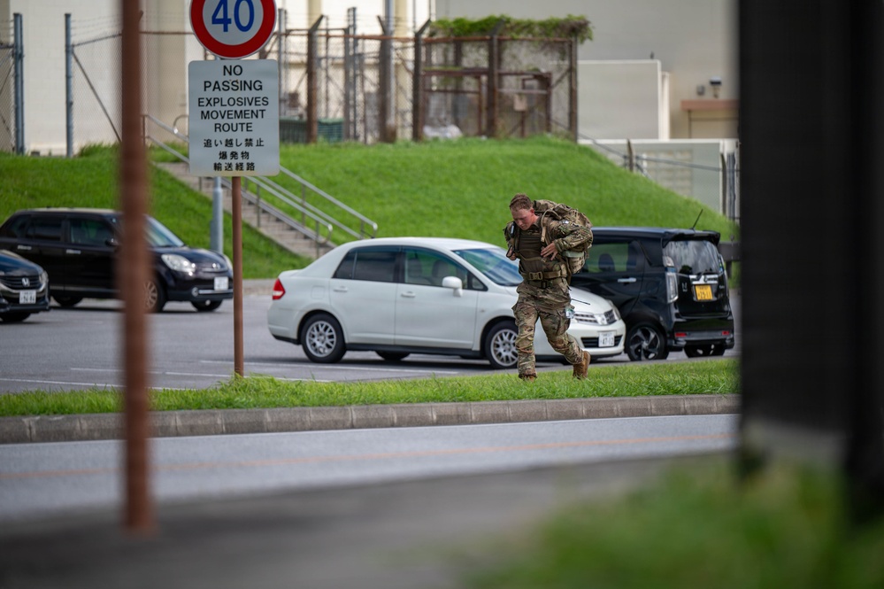 A military service member in full combat gear and a loaded rucksack is running across a grassy median on what appears to be a U.S. military installation in Japan. The scene includes bilingual signage in English and Japanese, indicating an explosives movement route restriction. The setting suggests a training exercise or rapid response drill on a joint-use military base.