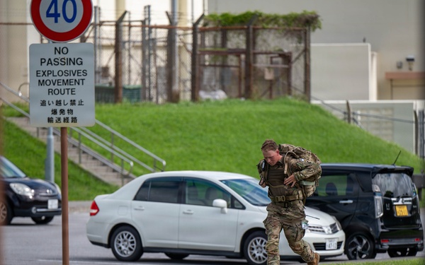 A military service member in full combat gear and a loaded rucksack is running across a grassy median on what appears to be a U.S. military installation in Japan. The scene includes bilingual signage in English and Japanese, indicating an explosives movement route restriction. The setting suggests a training exercise or rapid response drill on a joint-use military base.