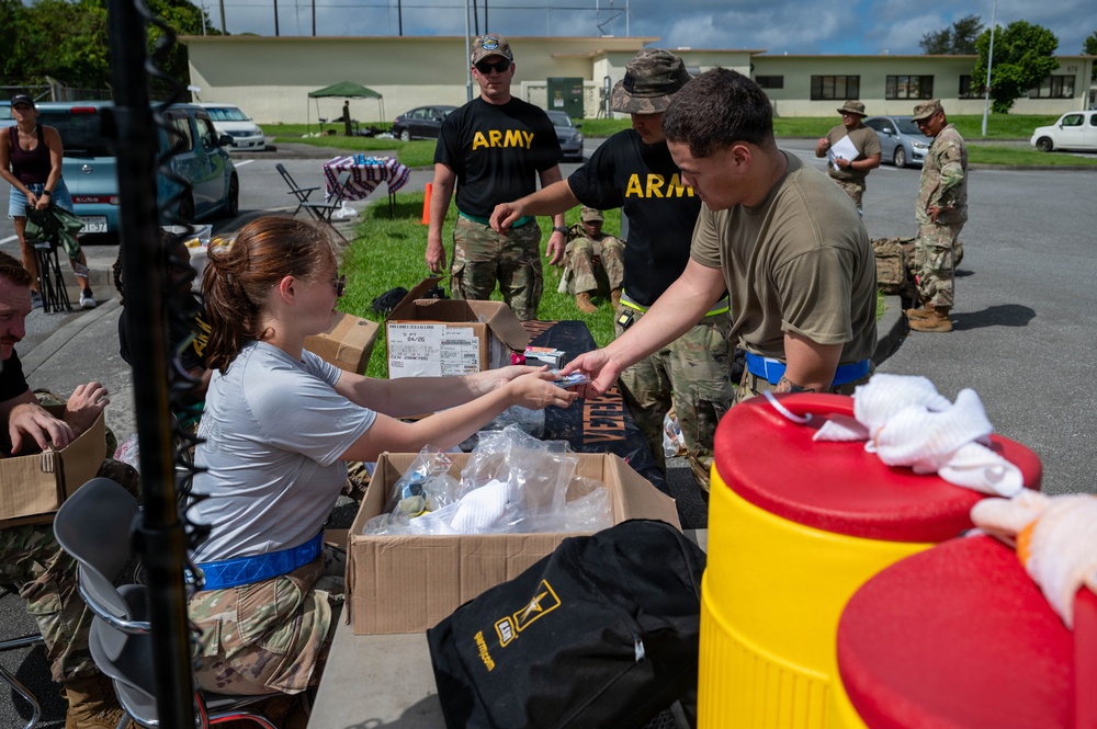 U.S. Army personnel are engaged in what appears to be a supply distribution or equipment staging operation outdoors on a military installation. A civilian or soldier in civilian attire is handing items from an open cardboard box to a soldier in combat uniform. The scene suggests a pre-deployment, training exercise, or humanitarian relief preparation activity.