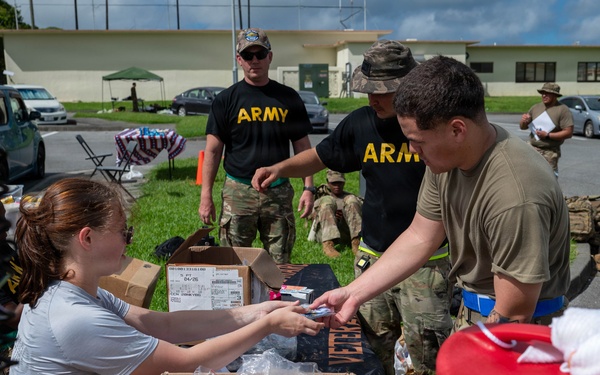 U.S. Army personnel are engaged in what appears to be a supply distribution or equipment staging operation outdoors on a military installation. A civilian or soldier in civilian attire is handing items from an open cardboard box to a soldier in combat uniform. The scene suggests a pre-deployment, training exercise, or humanitarian relief preparation activity.