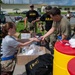 U.S. Army personnel are engaged in what appears to be a supply distribution or equipment staging operation outdoors on a military installation. A civilian or soldier in civilian attire is handing items from an open cardboard box to a soldier in combat uniform. The scene suggests a pre-deployment, training exercise, or humanitarian relief preparation activity.